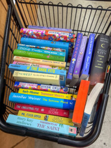 A top-down view of a black wire shopping cart filled with a colorful assortment of books from a variety of genres. Prominent visible titles include "Crescent City" by Sarah J. Maas, "Hello Stranger" by Katherine Center, and "The Summer Place" by Jennifer Weiner.