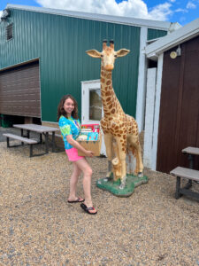 A young woman in a tie-dye shirt and pink shorts holds a box full of books while standing on a gravel lot next to a slightly smaller than life-sized statue of a giraffe.