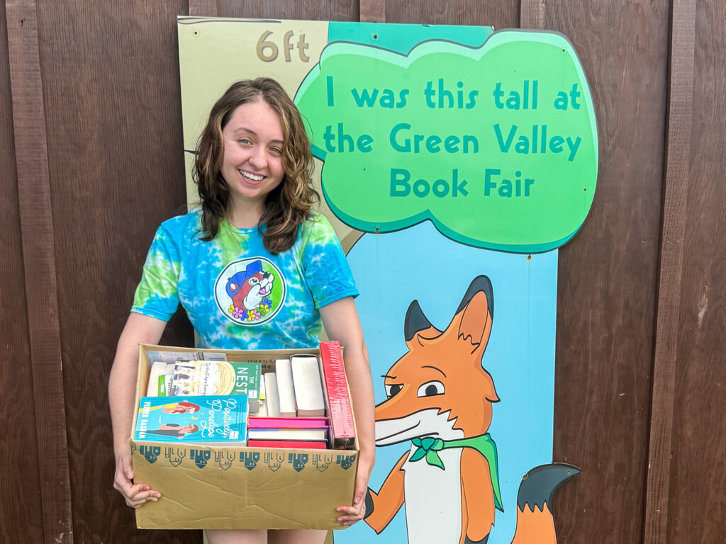 A smiling young woman in a blue and green tie-dye shirt holds a cardboard box brimming with books. She stands next to a sign with a cartoon fox that reads, "I was this tall at the Green Valley Book Fair."