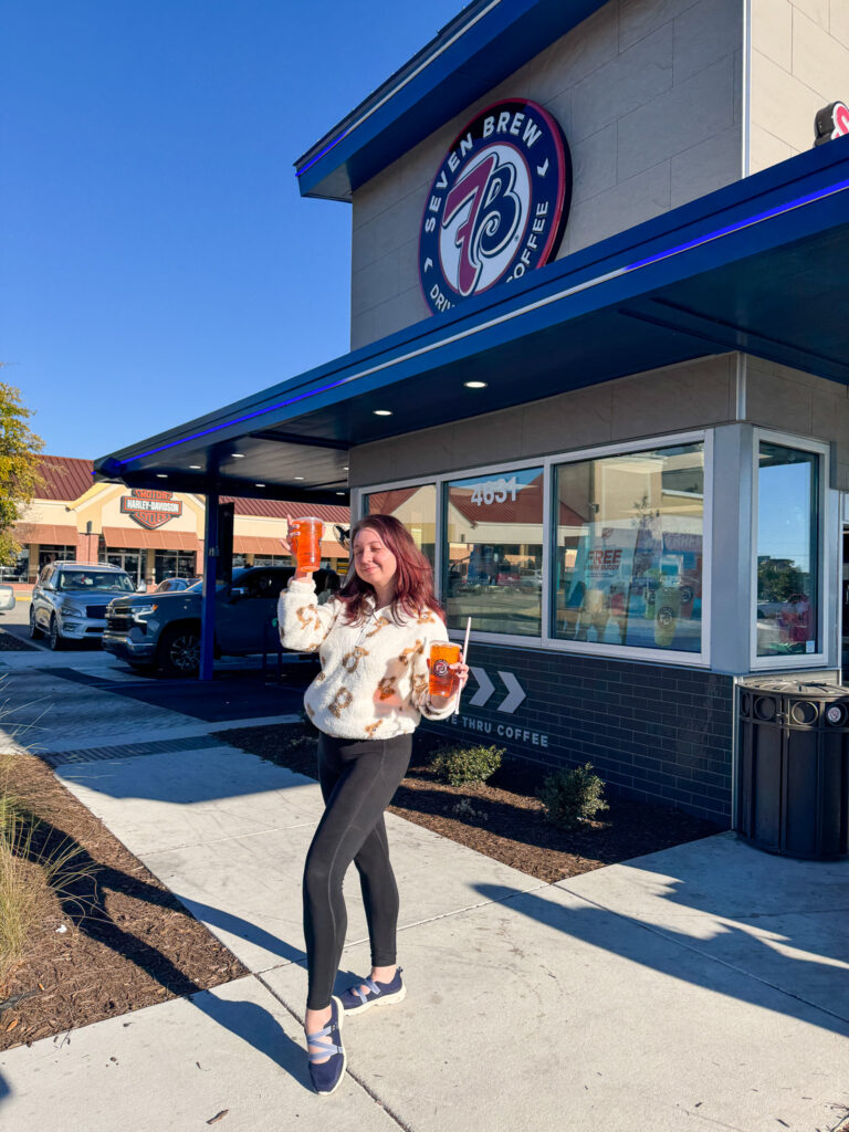 A woman with fuchsia hair poses in front of a Seven Brew Drive-Thru Coffee building, holding two large iced teas. She is wearing a cream-colored fuzzy quarter-zip with a tan teddy bear pattern, black leggings, and blue athletic slip-on shoes.