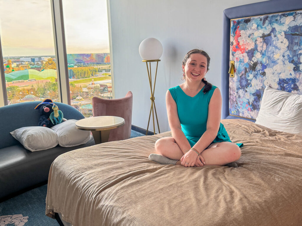 A young woman with brown hair in braids smiles while sitting cross-legged on a hotel bed with a light pink comforter. The room includes a tall window overlooking a theme park, a modern floor lamp, and a headboard decorated with a blue and pink abstract painting. A Captain Cacao plush and Stormfly plush sit on the lounge sofa near the window.