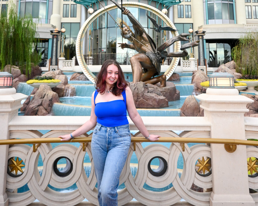 A smiling woman with fuchsia hair stands on a bridge in front of a grand, multi-tiered waterfall and a large golden statue of a celestial archer at Universal’s Epic Universe. She is wearing a vibrant blue sleeveless bodysuit, light-wash jeans with intricate rhinestone detailing, and a delicate silver necklace.