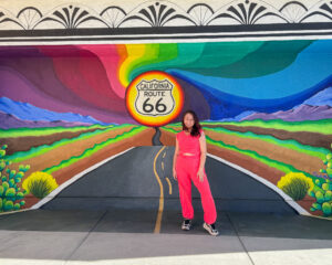 A wide-angle shot of a woman in a hot pink lounge set standing before a colorful, stylized Route 66 mural that depicts a road stretching toward a bright yellow sun.