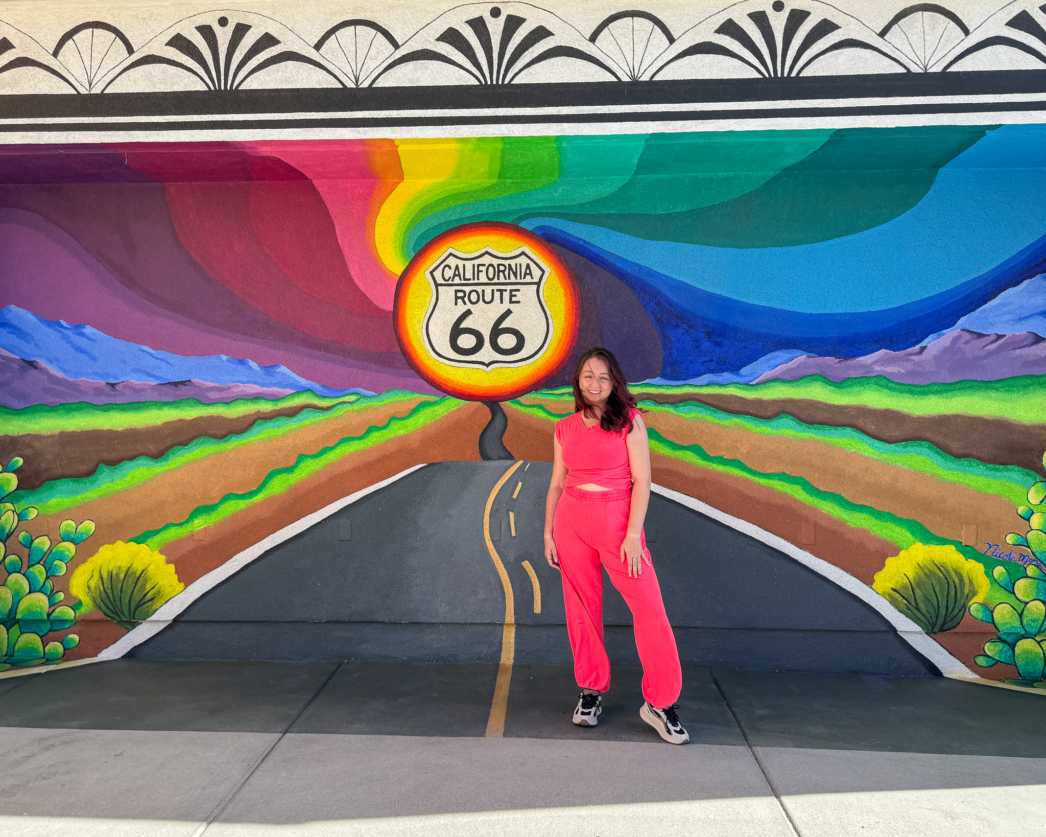 A wide-angle shot of a woman in a hot pink lounge set standing before a colorful, stylized Route 66 mural that depicts a road stretching toward a bright yellow sun.