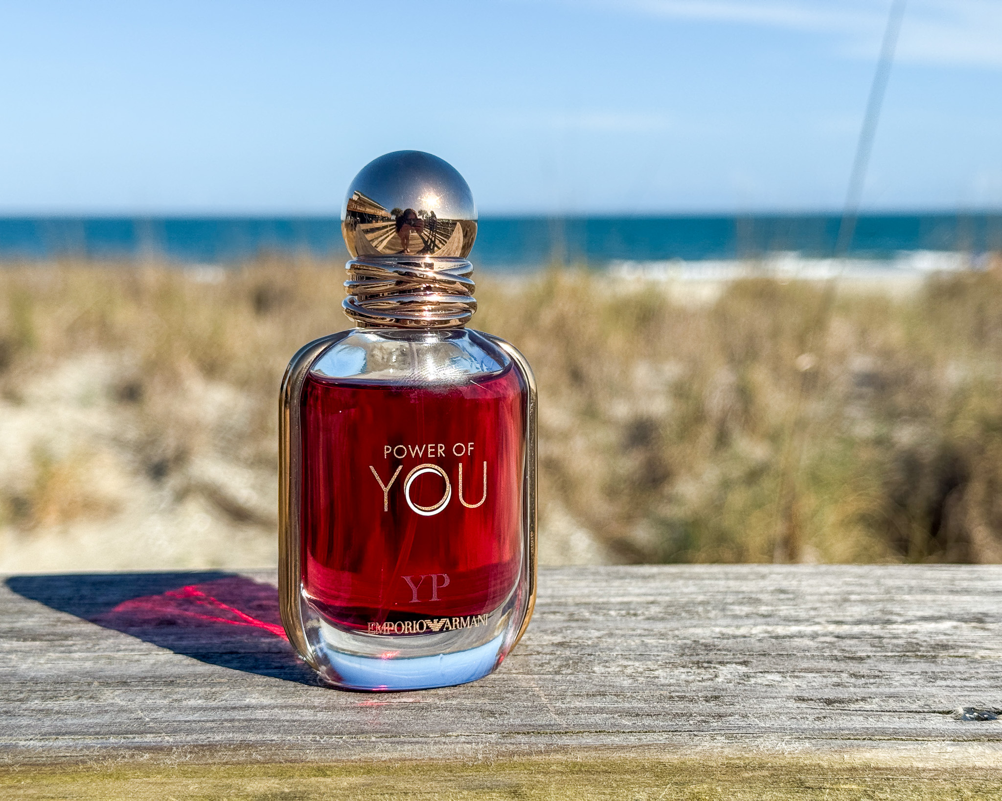 A bottle of Emporio Armani Power of You fragrance resting on a weathered wooden railing with a blurred background of a sandy beach, seagrass, and the blue ocean under a clear sky.
