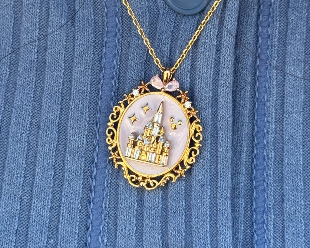 A detailed macro shot of an ornate oval locket necklace from Girls Crew, resting against a blue ribbed knit sweater. The 18k gold-plated locket features a pale, pearlescent pink background behind a raised, glittering Cinderella Castle encrusted with clear cubic zirconia crystals. The decorative gold border is framed with swirling filigree, tiny stars, and a small pink gemstone bow at the very top.