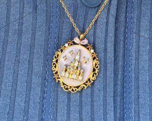 A detailed macro shot of an ornate oval locket necklace from Girls Crew, resting against a blue ribbed knit sweater. The 18k gold-plated locket features a pale, pearlescent pink background behind a raised, glittering Cinderella Castle encrusted with clear cubic zirconia crystals. The decorative gold border is framed with swirling filigree, tiny stars, and a small pink gemstone bow at the very top.