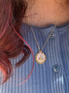 A medium close-up shot showing how the delicate gold Girls Crew Disney castle locket hangs on a dainty chain. The necklace is worn against a blue ribbed button-down sweater with dark blue buttons. Waves of long, dark hair with subtle reddish-pink highlights frame the left side of the shot.