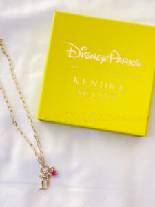 A flatlay featuring a bright yellow Disney Parks x Kendra Scott gift box on a white fabric background. Laid next to the box is a high-shine gold link chain necklace featuring a cluster of two distinct charms: a classic script Disneyland "D" logo, and a vibrant pink magenta stone charm cut into the signature Kendra Scott shape with a Mickey ear frame.