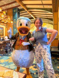 Woman in a blue short-sleeve top and blue floral jeans smiling arm-in-arm with Donald Duck dressed in his outdoor adventure scout vest and cap inside Storytellers Cafe.