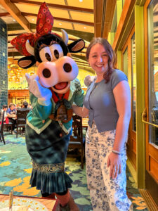 Woman posing with Clarabelle Cow, who wears a green patterned dress, sweater, and red floral bow, inside Storytellers Cafe.