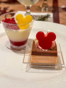 Two individual Disney dessert portions on a plate: a layered berry parfait cup topped with a yellow Mickey emblem and a square cake topped with pink cream and a red Mickey emblem.